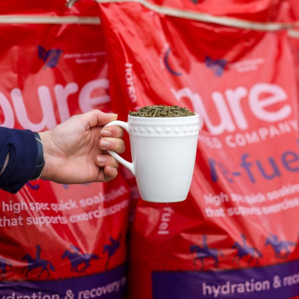 A hand holding a white mug filled with Pure Feed Refuel pellets, with red Refuel feed bags in the background.