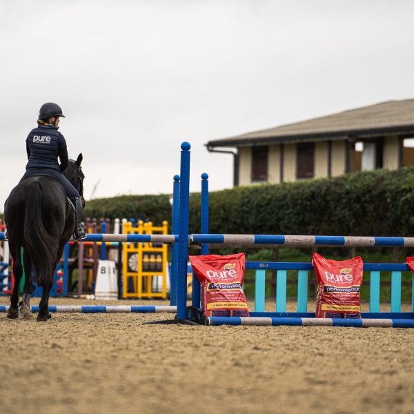 Pure Feed Performance – High-Energy Feed for Sport & Competition A rider on horseback in an arena with Pure Feed Performance horse feed bags placed along the showjumping course.